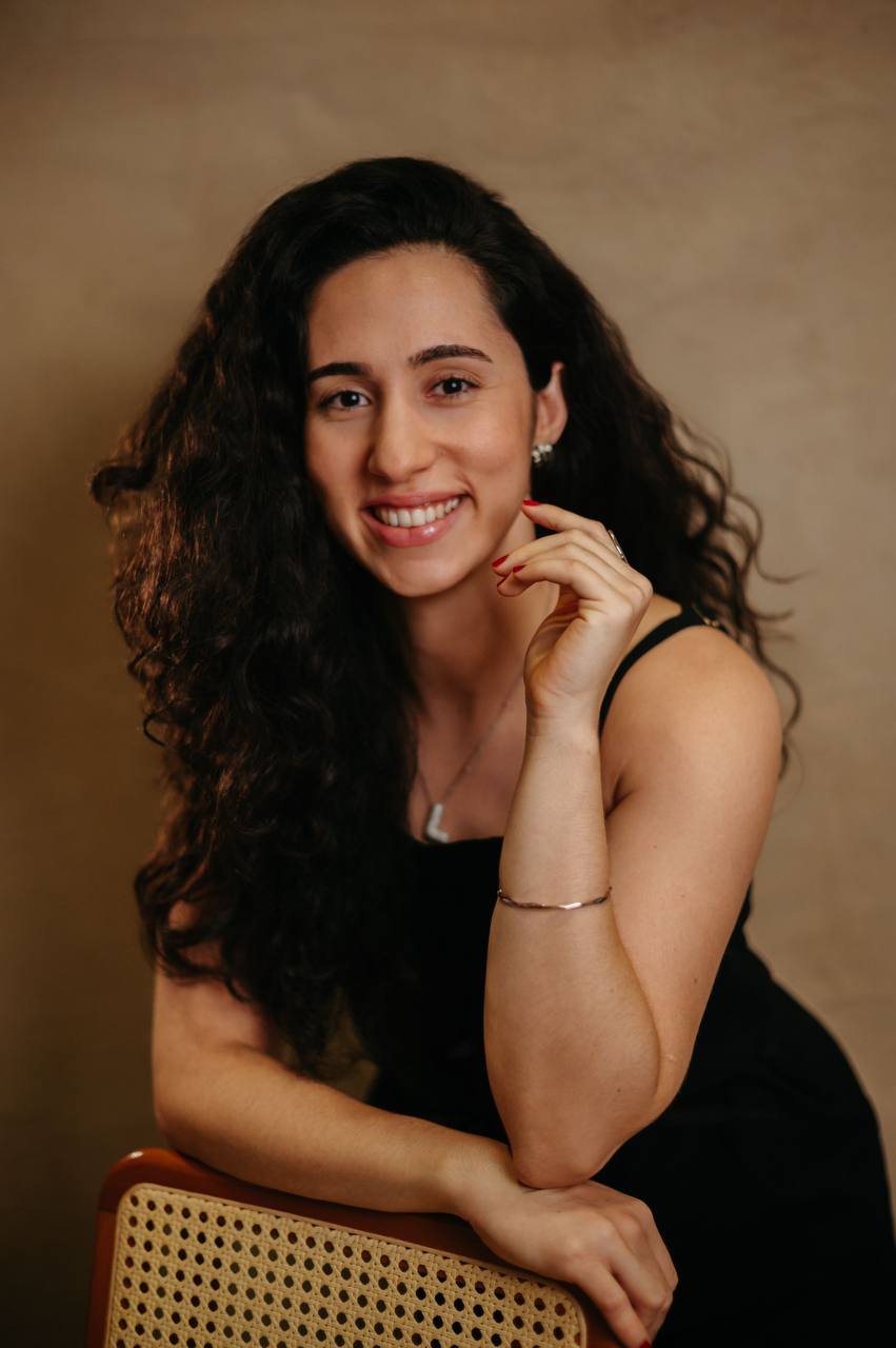 Nutricionista Larissa sorrindo em um retrato profissional, com roupa preta e fundo em tons quentes.